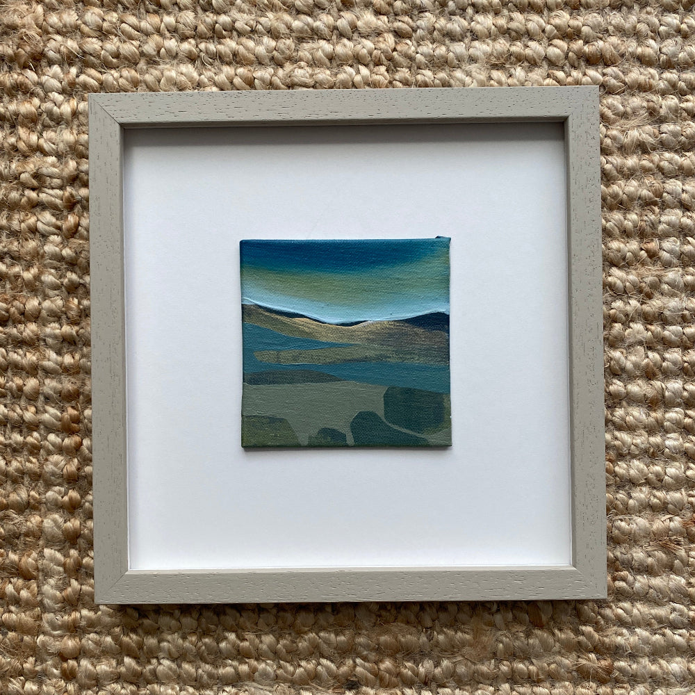 Framed artwork of a landscape with blue and green tones on a textured beige surface.