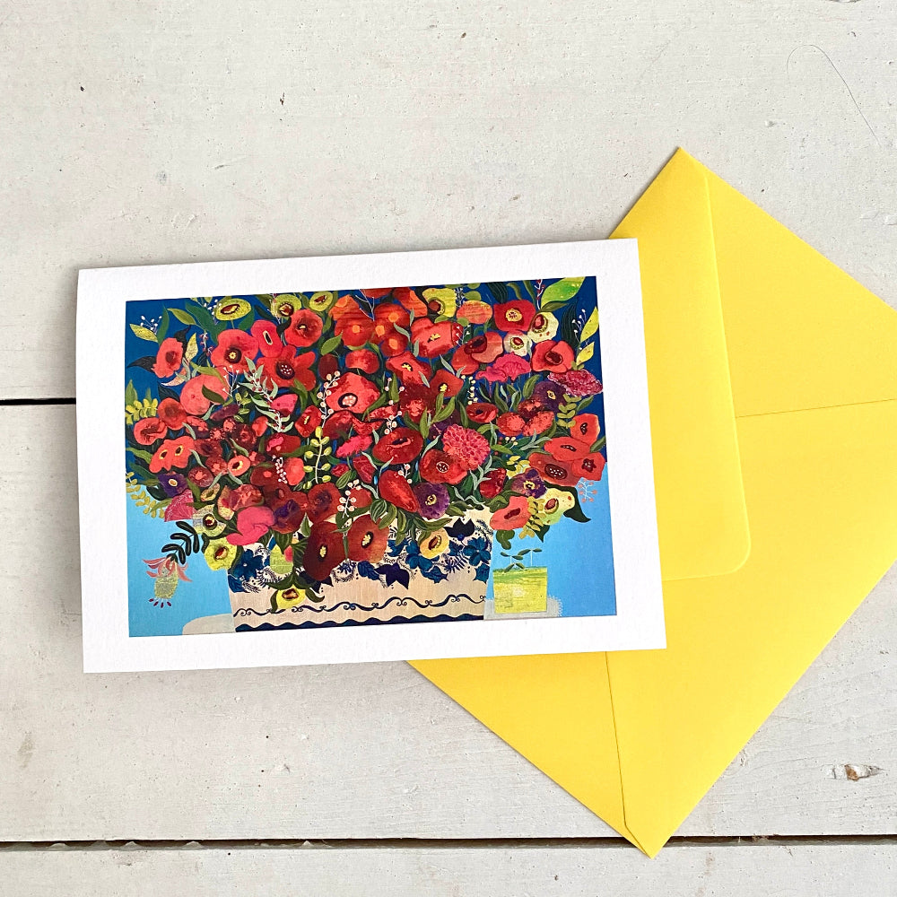 A square greeting card with a colorful floral illustration in a vase, placed on a light wood surface with a bright yellow envelope behind it.