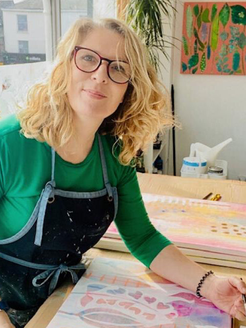 A woman with blonde hair and glasses, wearing a green shirt and a dark paint-splattered apron, smiling while looking at a colorful painting she is working on at a table.
