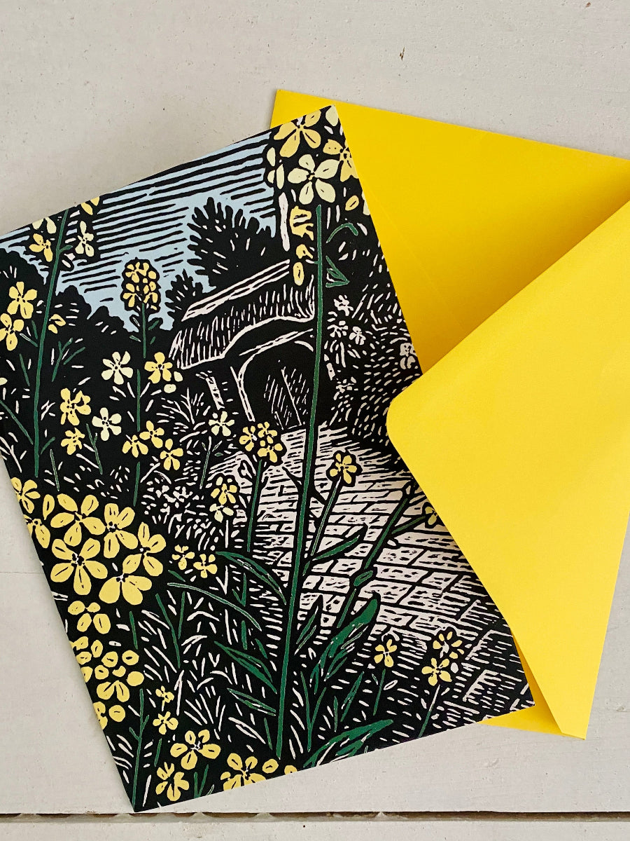 A square printed card or envelope with a black-and-white linocut-style floral and architectural illustration, layered over a bright yellow envelope on a light neutral surface.