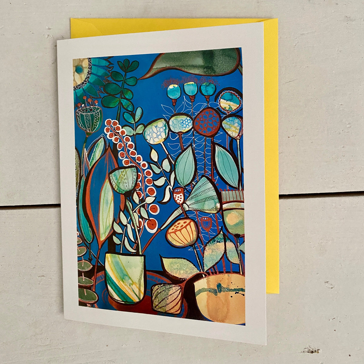 A matted art card featuring a colourful botanical illustration on a deep blue background, displayed on a light grey wooden surface with a yellow envelope behind it.