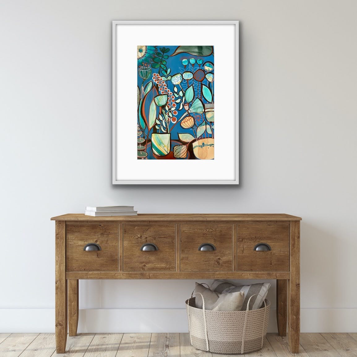 A framed abstract art print with a white mat and silver frame hangs on a light gray wall above a rustic wooden console table. The artwork features a colorful abstract design with blue, green, orange, and beige shapes.