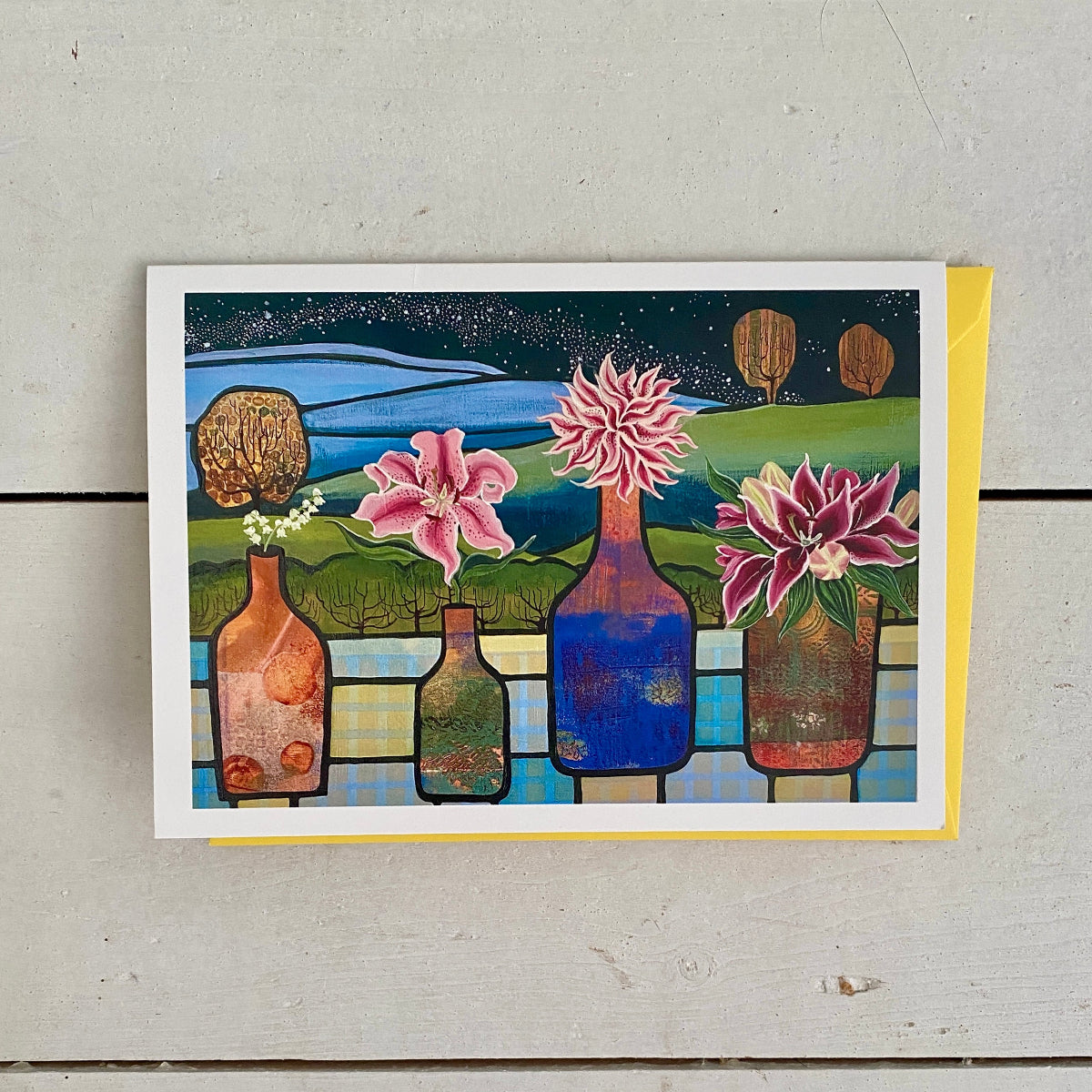 A square art print or greeting card with a colorful illustration of flowers in bottles against a landscape background, displayed on a light gray wooden surface with a yellow envelope behind it.