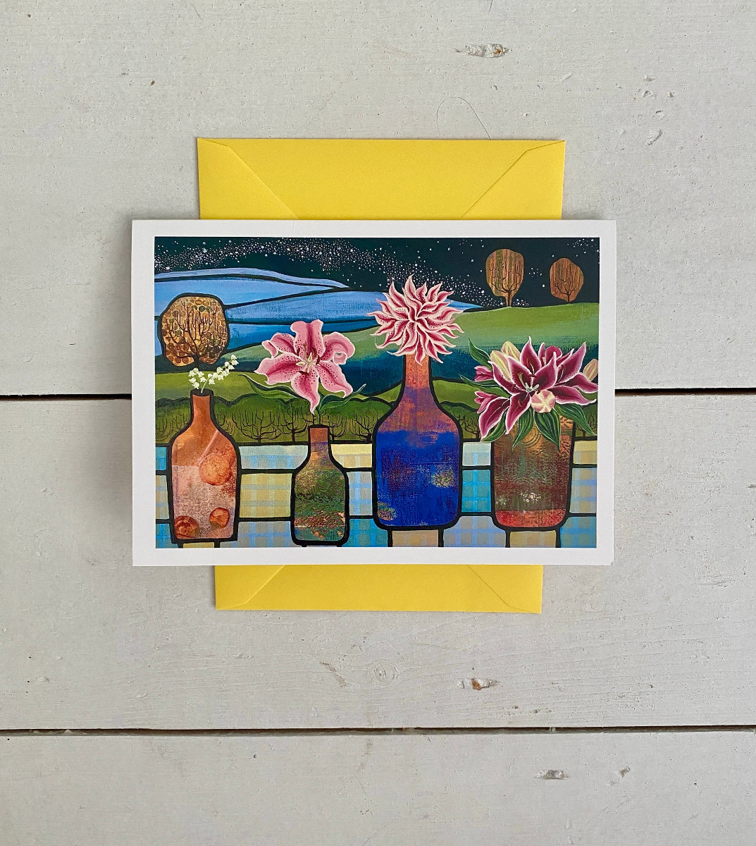 A square art print or greeting card with a colorful illustration of flowers in bottles against a landscape background, placed on top of a yellow envelope over a light wood surface.