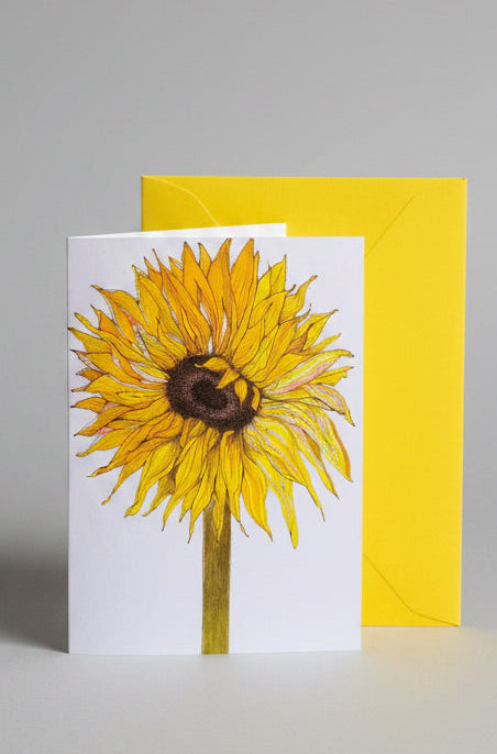 A white greeting card featuring a detailed illustration of a yellow sunflower, standing in front of a bright yellow envelope on a neutral pale gray background.