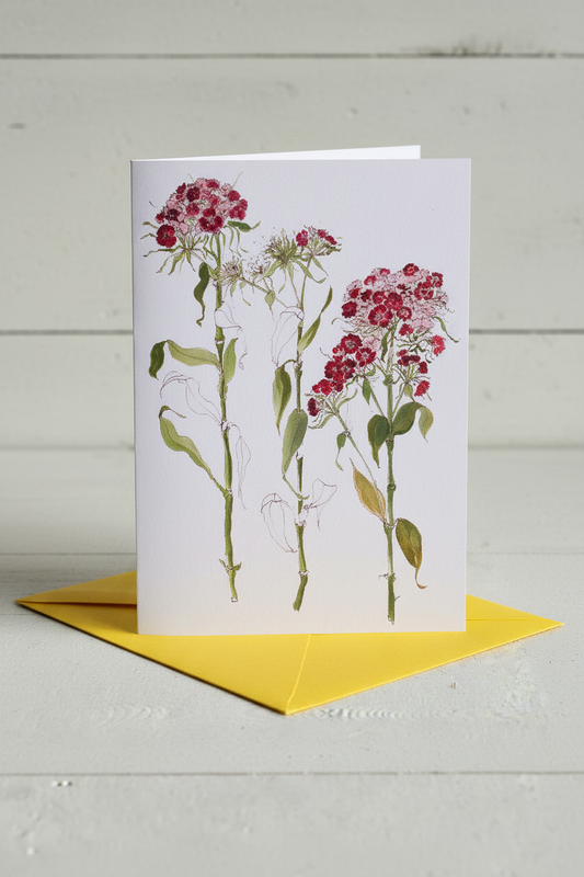 A white greeting card featuring a botanical illustration of three pink and red flower clusters with green stems and leaves, displayed upright on a bright yellow envelope against a light gray wood plank background.