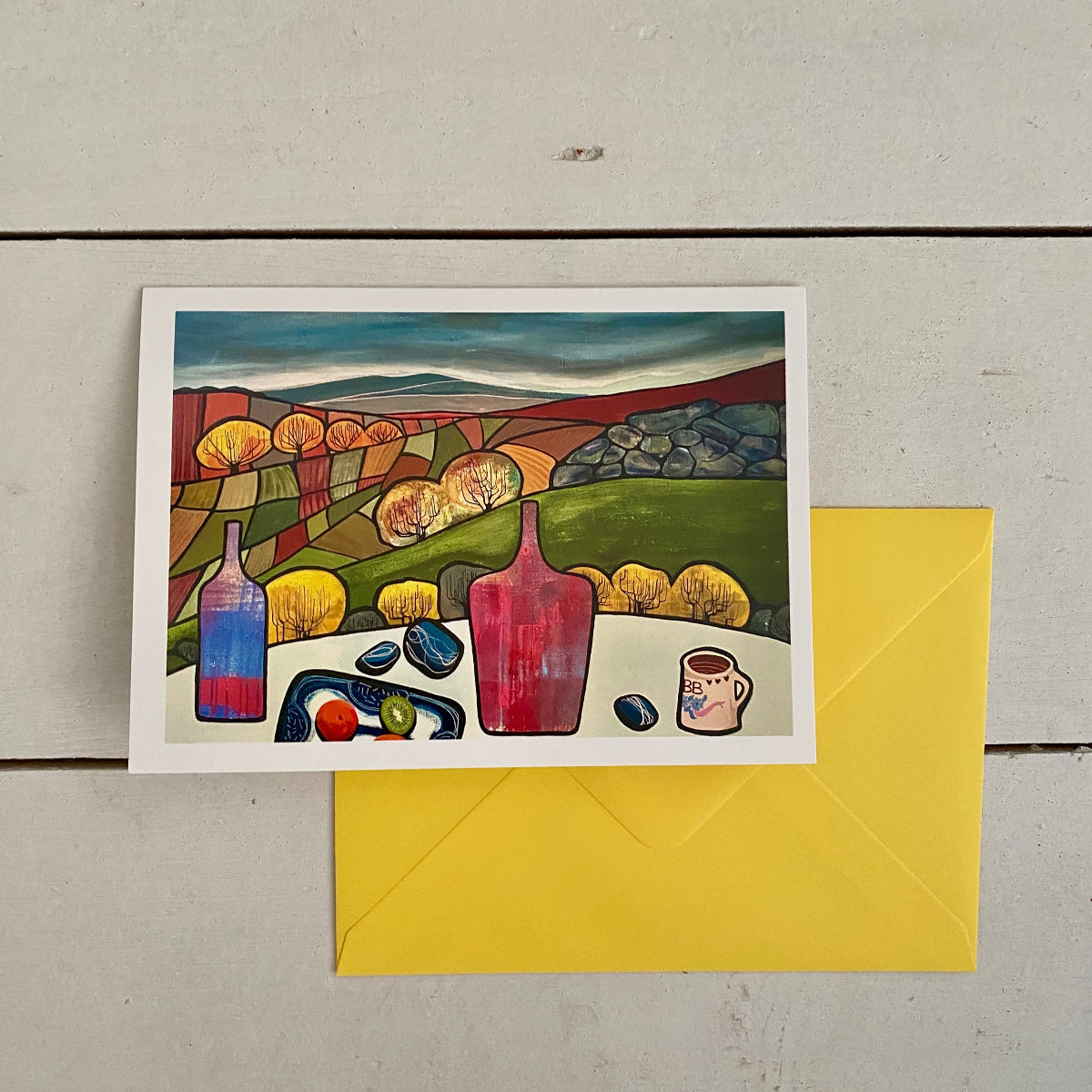 A square greetings card with a white border showing a colourful still life landscape painting, placed on top of a bright yellow envelope on a light gray wood background.