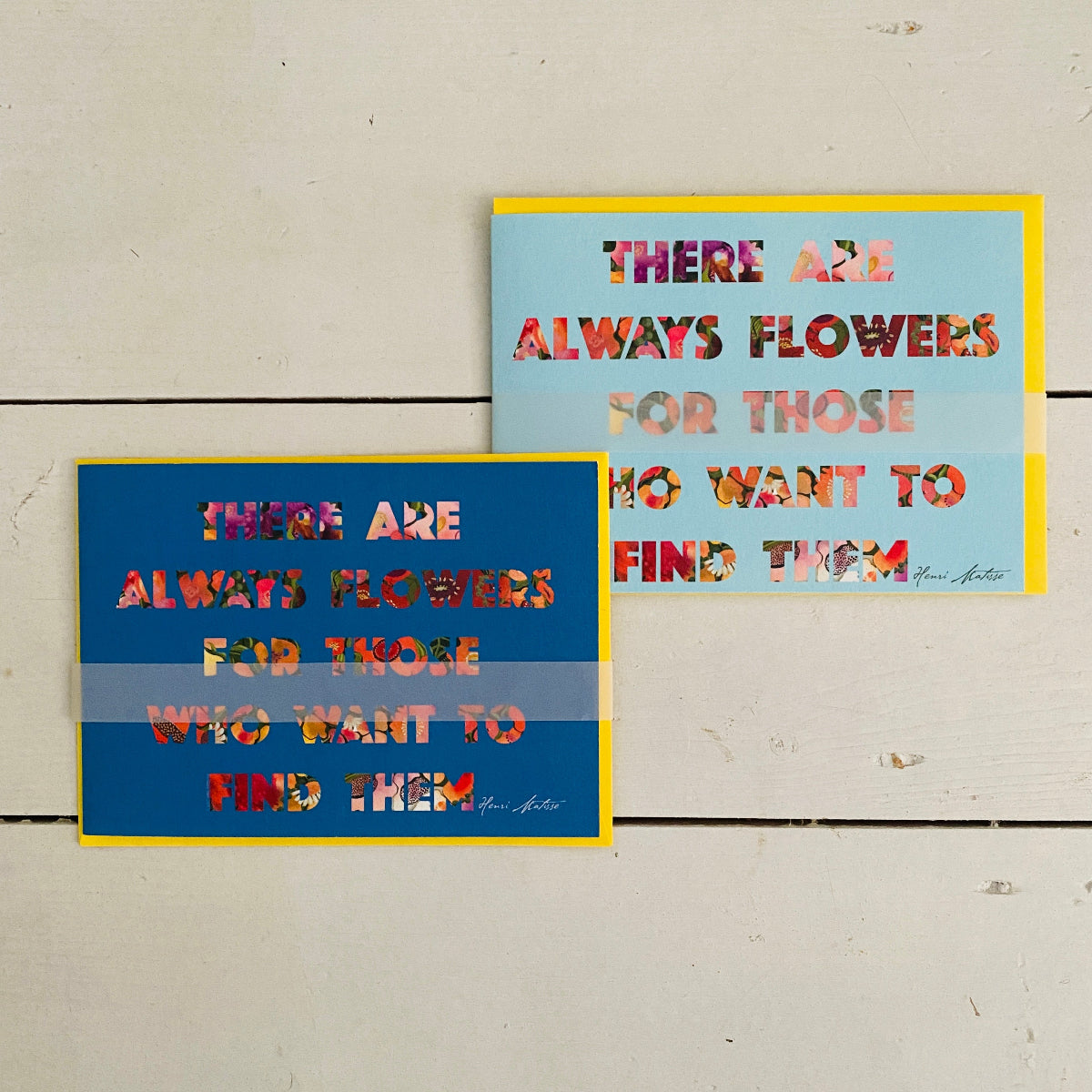 Two rectangular landscaped format art prints with motivational text are displayed on a white wooden background. The top print has a light blue background with yellow borders and multicolored floral lettering.