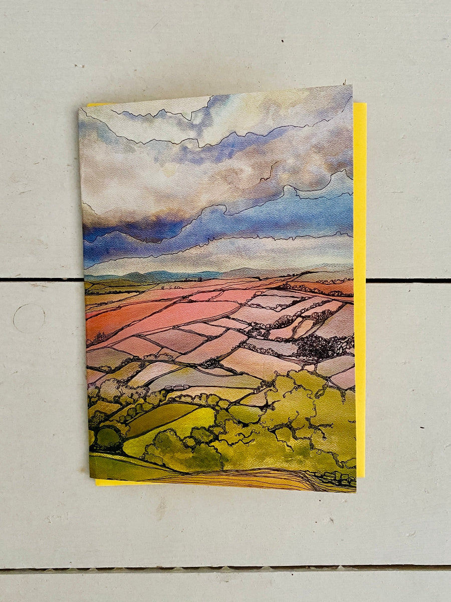 A vertical watercolor landscape art print depicting a patchwork of green and pink fields under a cloudy blue sky, displayed on a light-colored wooden surface.