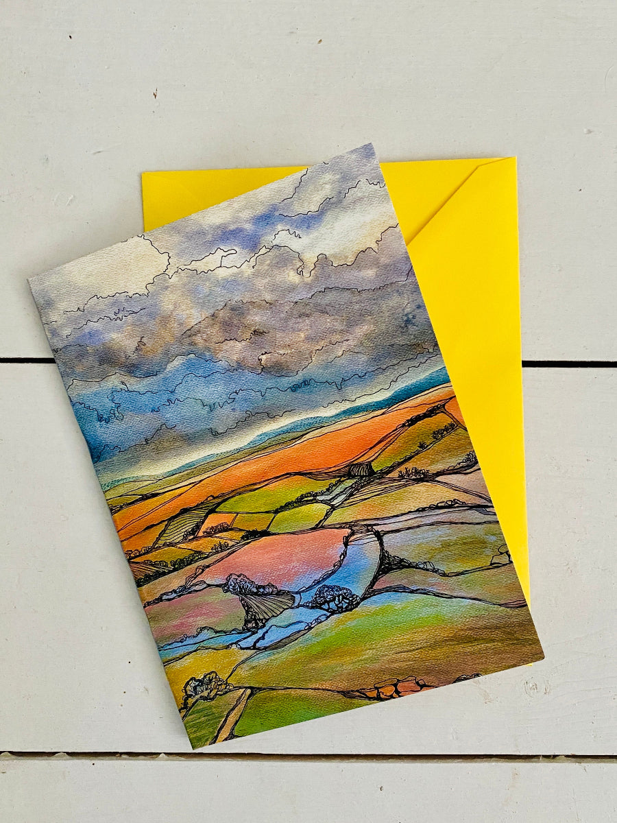 A landscape art print or postcard featuring a colorful hand-drawn countryside scene with fields, hills, and a dramatic cloudy sky, placed over a bright yellow envelope on a light wood surface.