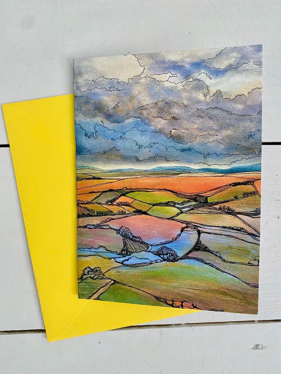 A watercolor landscape art print of rolling green and orange fields under a cloudy blue sky, displayed on a white wooden surface next to a bright yellow envelope.
