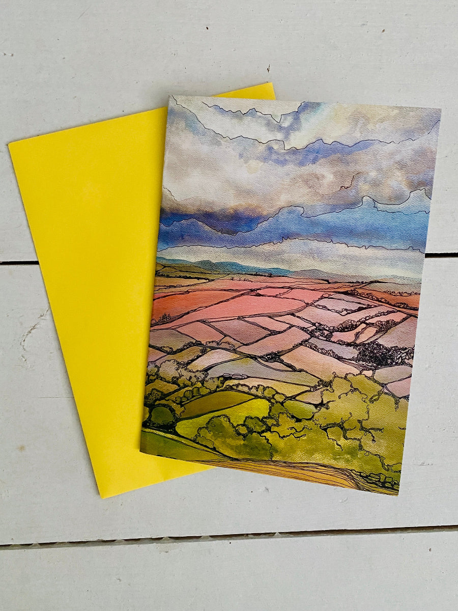 A watercolor landscape art print depicting a patchwork field under a cloudy sky, displayed on a white wooden surface next to a bright yellow envelope.