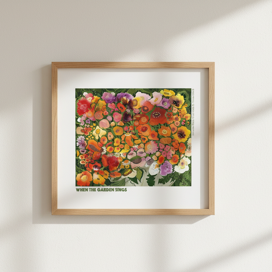 A framed art print featuring a colorful illustration of wildflowers with the text 'WHEN THE GARDEN BLOOMS' at the bottom, displayed on a clean white wall.