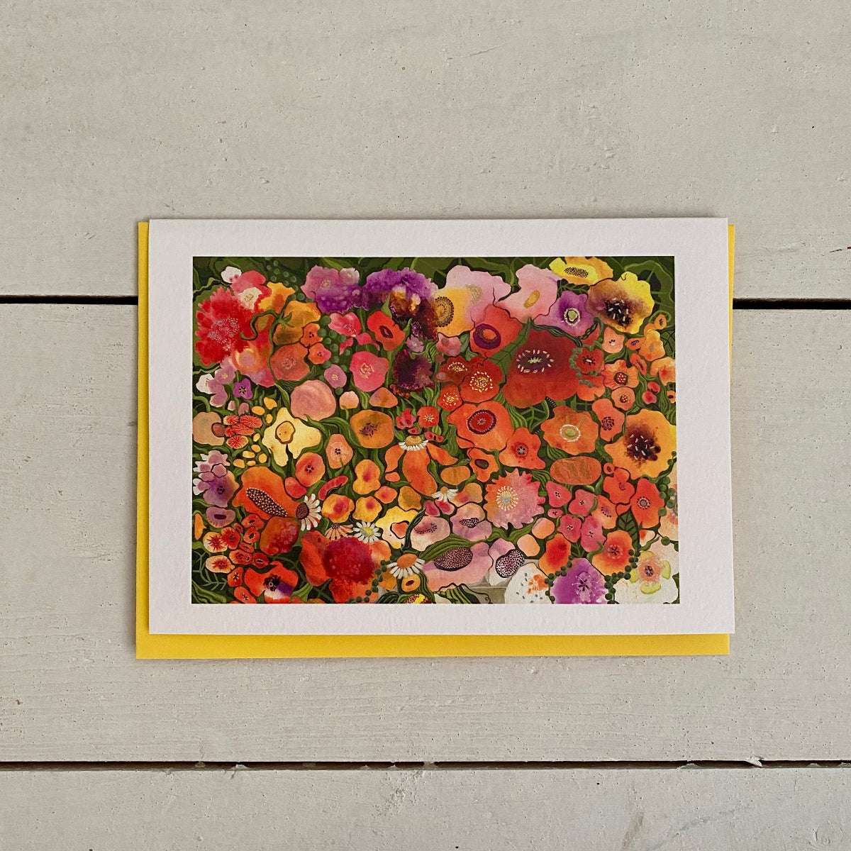 A rectangular art print or card featuring a dense, colorful floral illustration, displayed on a light gray wooden surface with a yellow backing card visible around the white border.