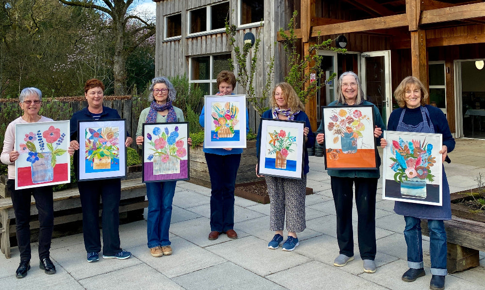 Group of women holding paintings outdoors in front of a wooden garden building. RHS Rosemoor classroom.