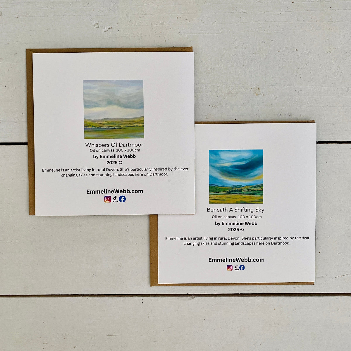 Two framed art prints with kraft paper borders are arranged on a white wood background. The larger print on the left reads 'Whispers Of Dartmoor' and the smaller print on the right reads 'Beneath A Shifting Sky', both featuring landscape photography by Emmeline Webb.