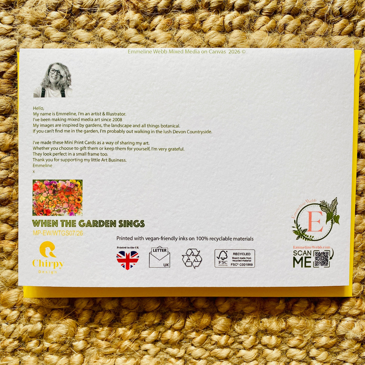A printed mini postcard card laid flat on a textured woven straw surface. The card has a white background with black text, a small portrait photo, a colourful floral illustration, and several logos and icons including Chirpy Design, When the Garden Sings, and Scan Me.