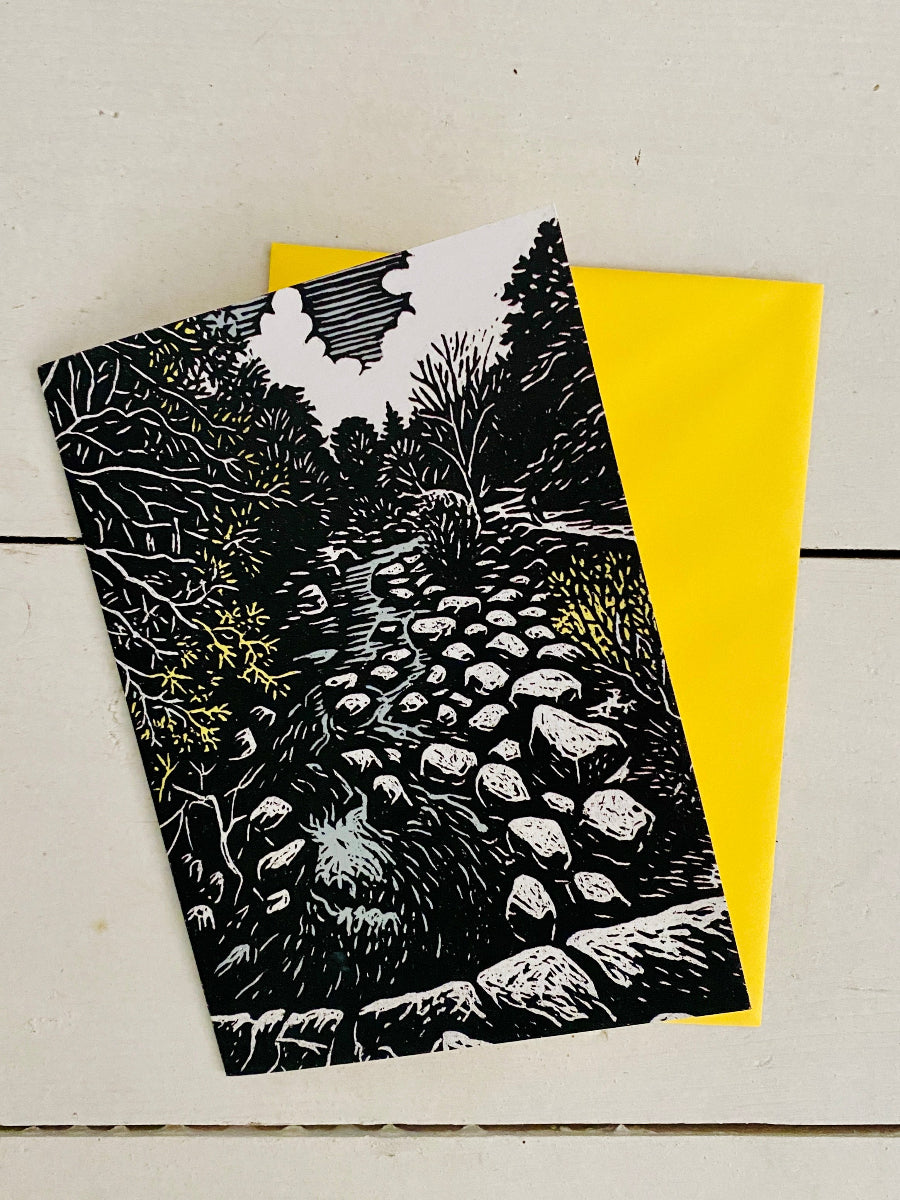 A black and white linocut-style art print depicting a rocky forest path, layered over a bright yellow envelope on a light wood background.