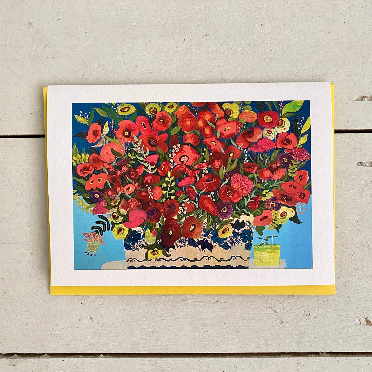A square art print or greeting card featuring a colorful floral illustration with red poppies and other flowers in a blue vase, photographed on a light weathered wood surface with a yellow card backing visible.