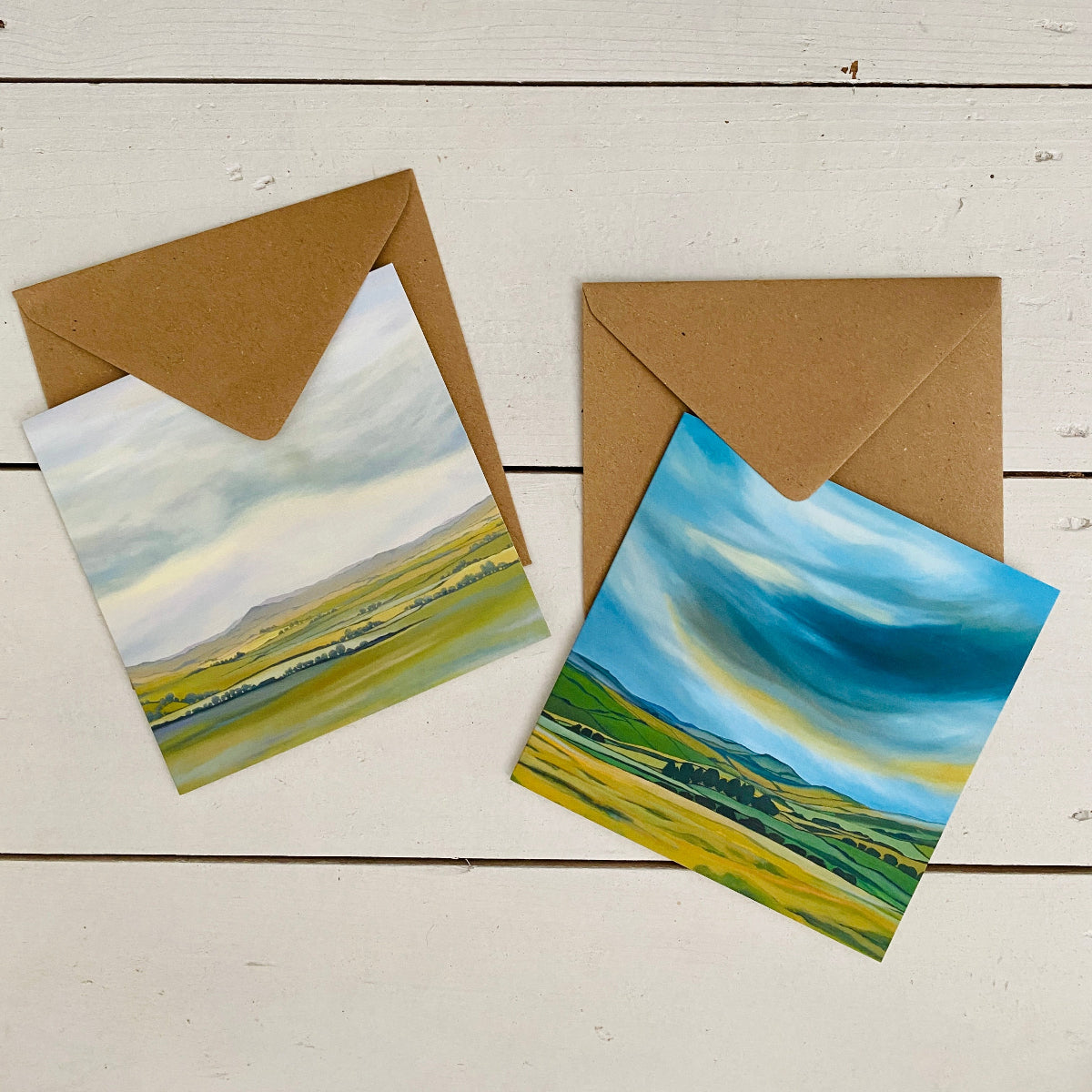 Two square greeting cards with brown kraft envelopes, laid flat on a white wood background. One card shows a soft landscape in muted green, yellow, and gray tones, and the other shows a vivid landscape with blue sky, green fields, and yellow hills.
