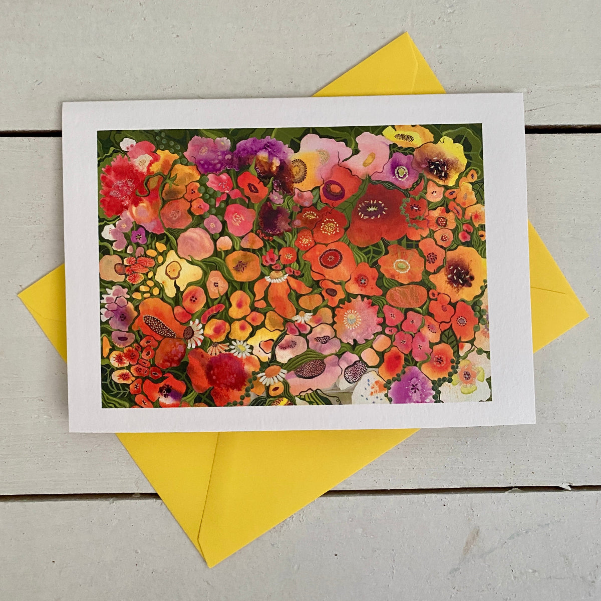 A square art print featuring a dense, colorful floral illustration, displayed on top of two yellow envelopes on a light gray wooden surface.
